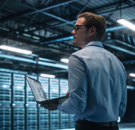 Man holding a laptop in a data center, looking over rows of server racks in a high-tech environment.