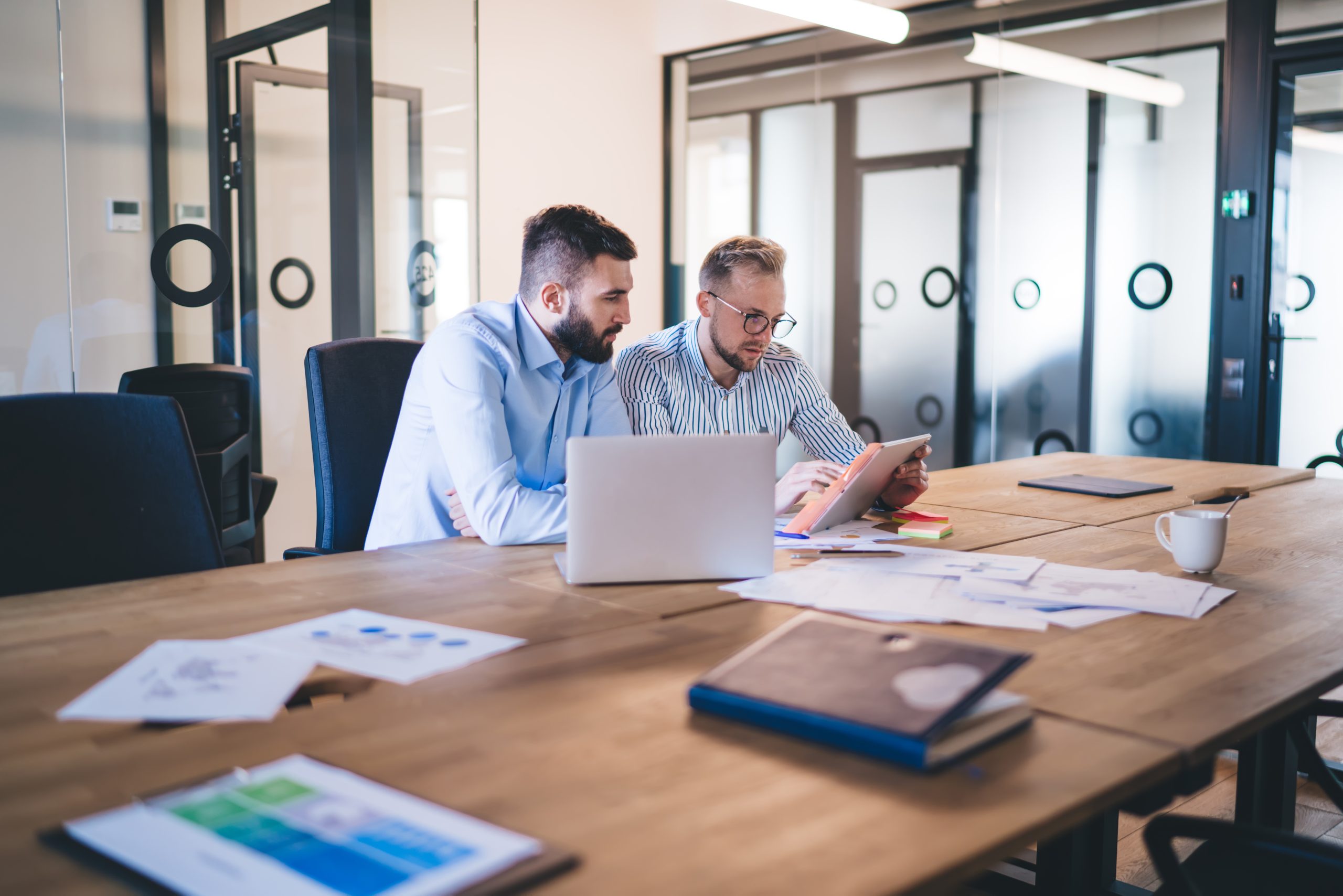 Smart caucasian male colleagues concentrated on reading report talking to each other for analyzing data, skilled professional men employees cooperating on project together sitting at table in office