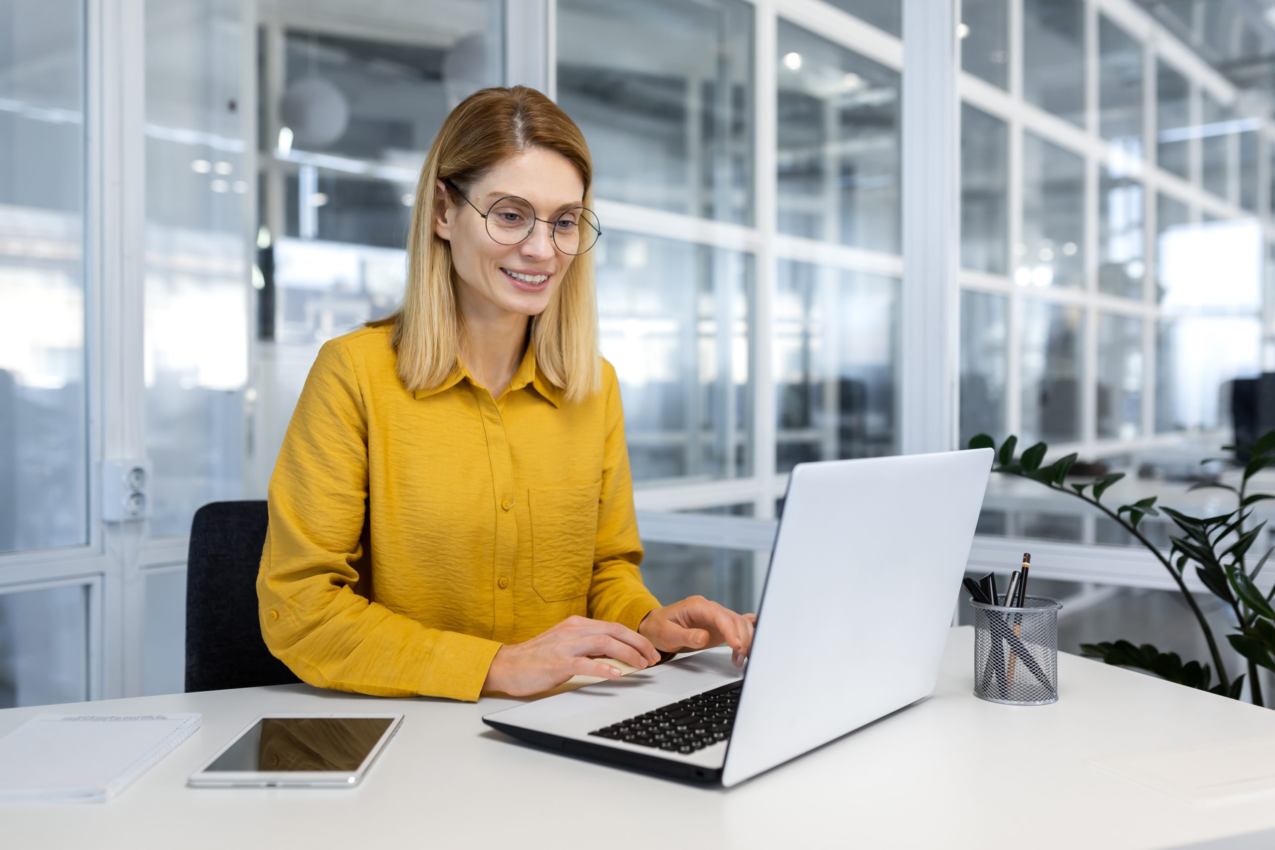Happy and successful business woman working inside office with laptop, female worker in yellow shirt and glasses typing on keyboard, woman satisfied with work and happy at workplace sitting at table