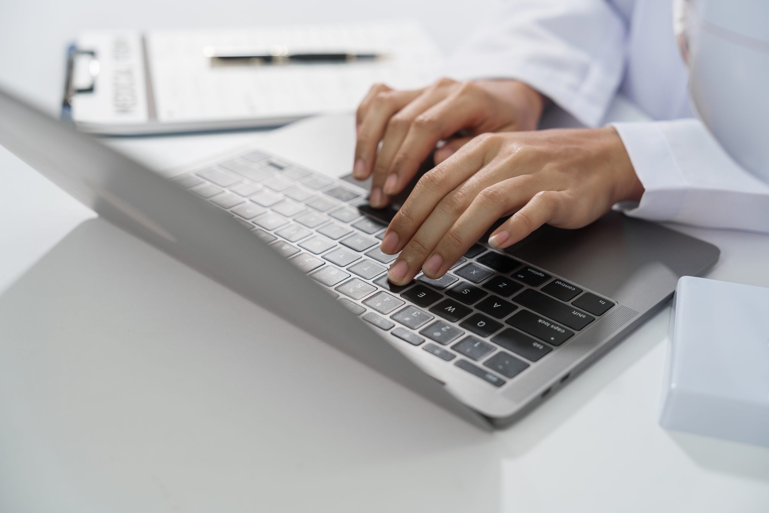 A doctor's hands are seen typing on a laptop keyboard with a stethoscope around the neck on white desk.