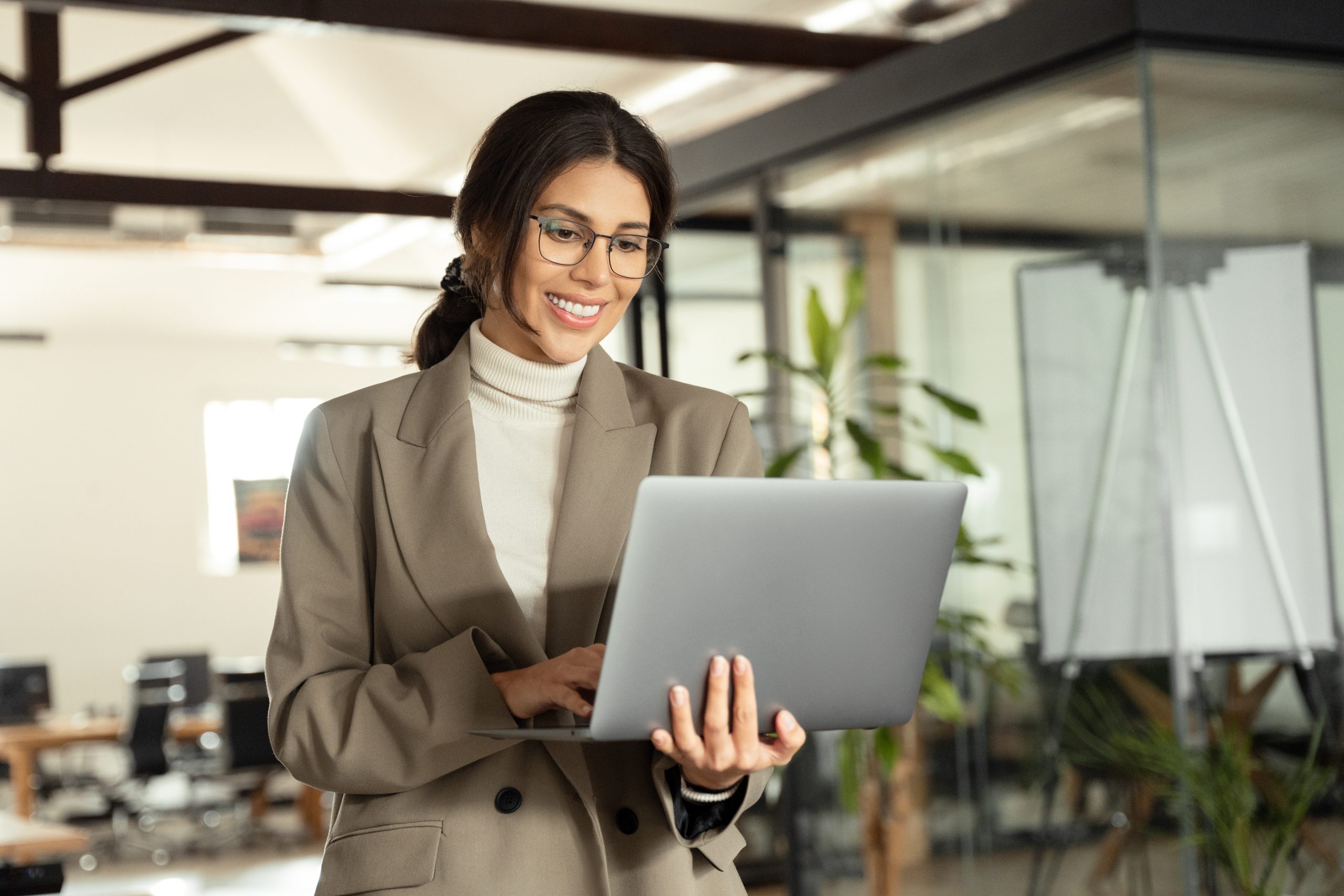 Smiling young businesswoman manager holding laptop pc using it fintech application standing at workplace in office. Latin Hispanic mature adult professional business woman working focused on computer