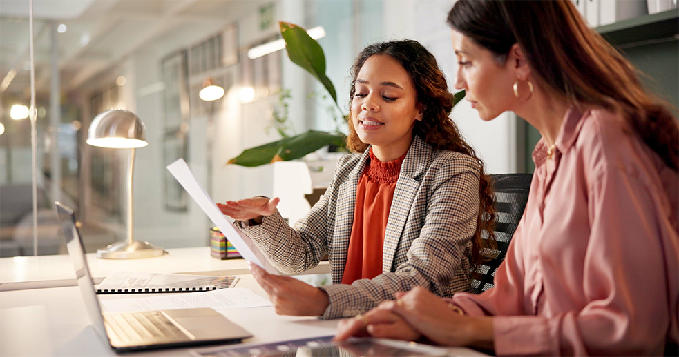Two women in a modern office reviewing a document together at a desk with a laptop, discussing and pointing at the paper under soft lighting.
